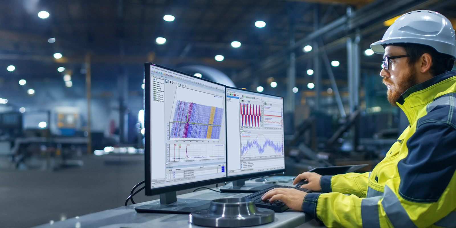 Person in safety gear working on two monitors with industrial data in a factory setting.