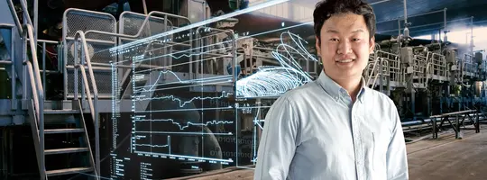 A man in a light blue shirt smiles in front of a factory setting, with a transparent overlay showing process monitoring data and graphs.